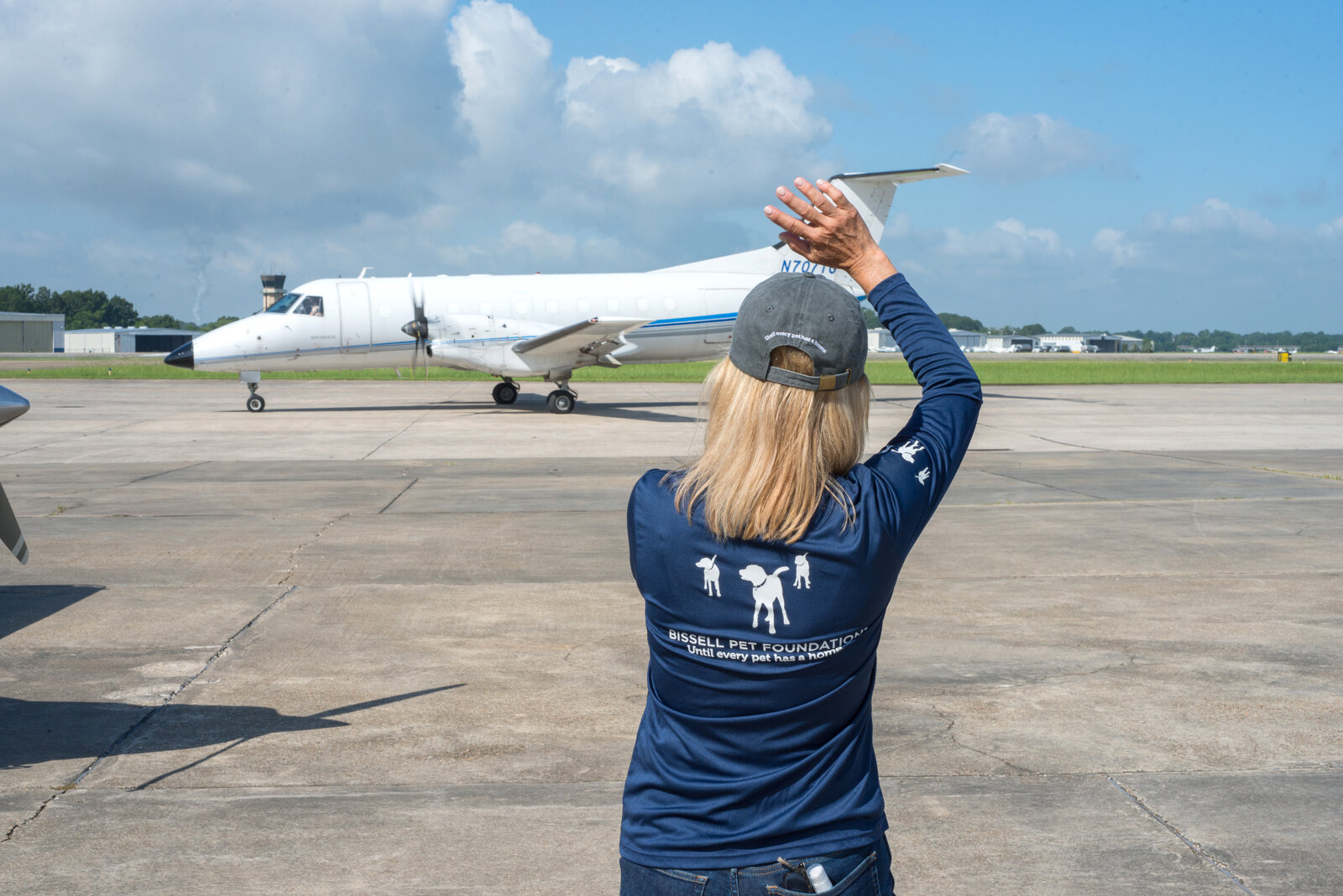A person wearing a Brishall Pet Foundation shirt stands on an airport tarmac, raising their arms to signal as a small white jet taxiing approaches on a sunny day with blue sky and scattered clouds.