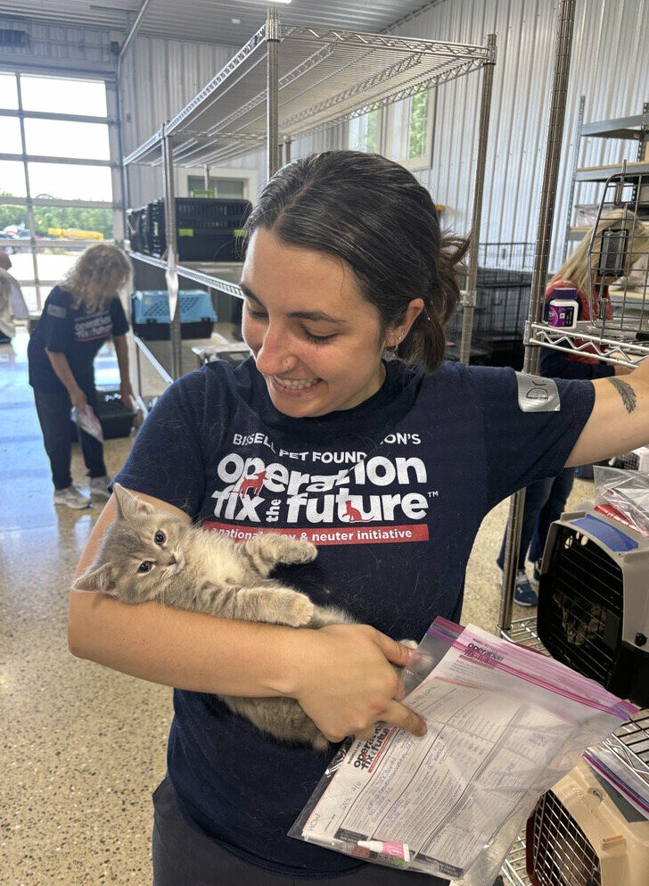 A smiling woman in a navy Operation Fix the Future shirt holds a gray tabby kitten while carrying paperwork inside an animal shelter. Other people and pet carriers are visible in the background.