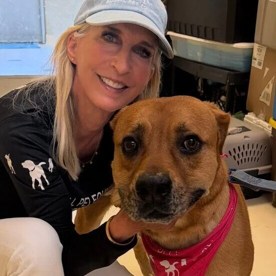 Smiling woman in a light blue cap crouches next to a brown dog wearing a red bandana indoors. The woman has her arm around the dog, and they both look at the camera.