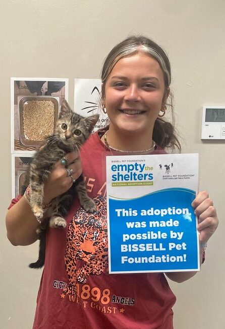 A smiling woman holds a small tabby kitten and a sign that reads, This adoption was made possible by BISSELL Pet Foundation! They stand indoors near a wall with cat-related posters.