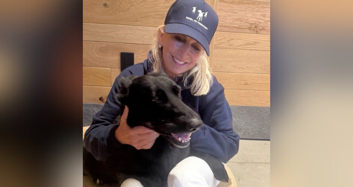 A woman in a navy blue cap and hoodie sits indoors, smiling as she hugs a happy black dog. They are in front of a wooden wall.