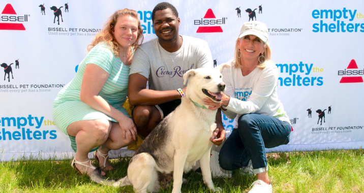 Three people kneel on grass, smiling and posing with a happy, large white and gray dog in front of an “empty the shelters” event backdrop with BISSELL Pet Foundation logos.