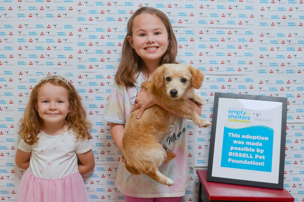 Two young girls smile at the camera; one holds a small tan dog. They stand in front of an “Empty the Shelters” backdrop next to a sign stating, “This adoption was made possible by BISSELL Pet Foundation!”.