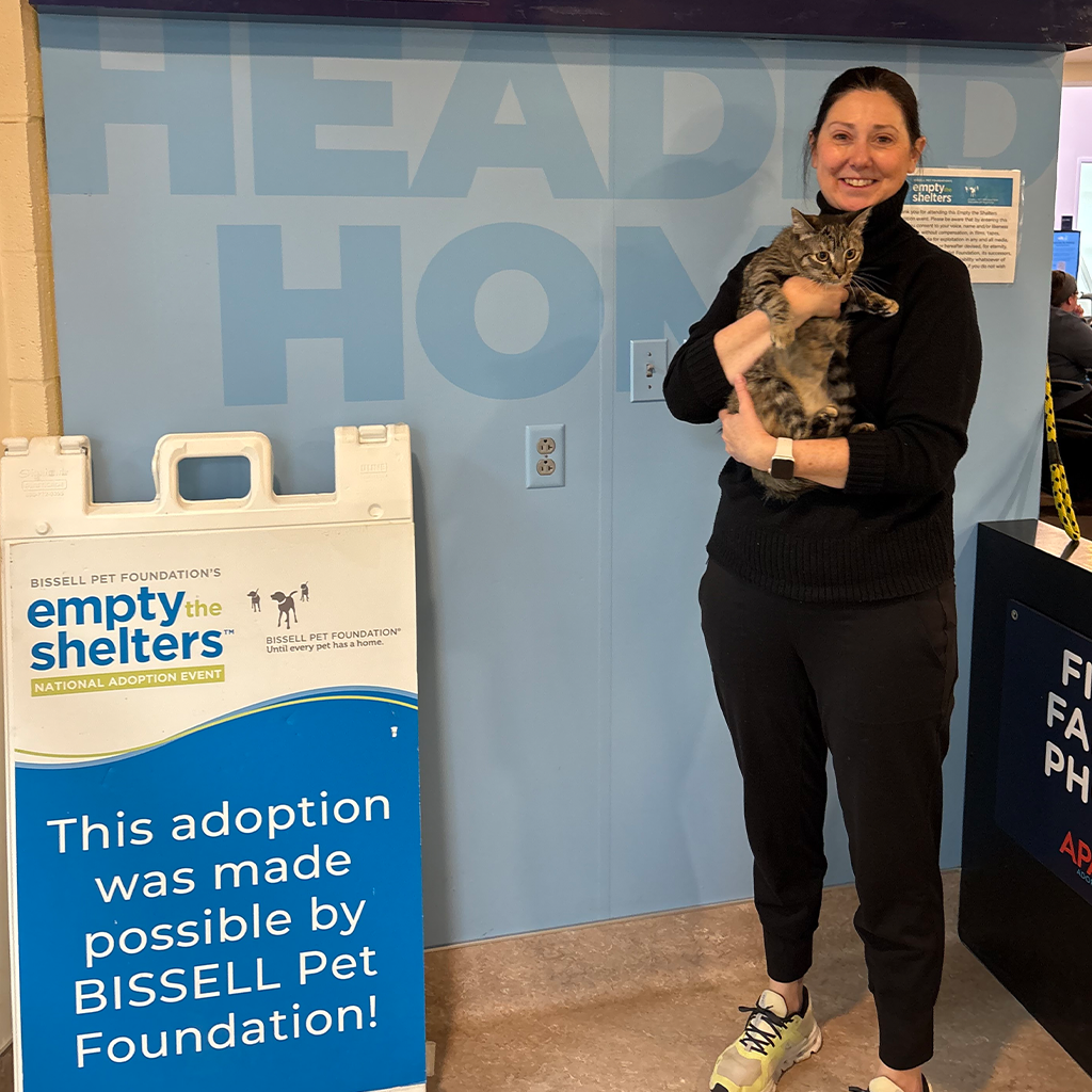 A smiling person in black clothes holds a tabby cat next to a sign reading, This adoption was made possible by BISSELL Pet Foundation! The sign promotes the empty the shelters national adoption event.