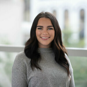 A woman with long, wavy dark hair and blue eyes is smiling at the camera. She is wearing a gray sweater and standing in front of a bright, blurred background with windows and greenery.