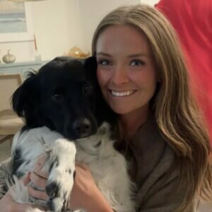 A smiling woman with long brown hair hugs a black and white dog indoors. The woman is facing the camera, and the dog’s front paws are up as it sits comfortably in her arms.