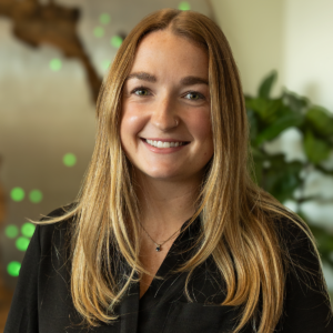 A young woman with long blonde hair, wearing a black shirt and a necklace, smiles at the camera. The background is softly blurred, featuring green plants and abstract shapes.