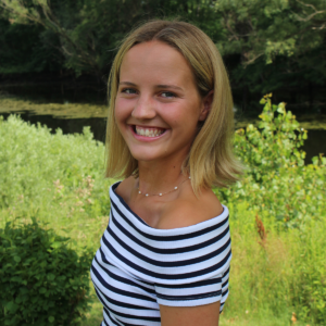 A young woman with blonde hair, wearing an off-the-shoulder black and white striped top, smiles while standing outside in a lush, green area with trees and a pond in the background.