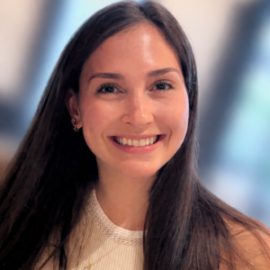 A woman with long brown hair and light skin smiles at the camera. She is wearing a sleeveless, light-colored top and small hoop earrings. The background is blurred, featuring soft blue and beige tones.