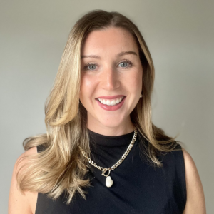 A woman with long, wavy blonde hair smiles at the camera. She is wearing a sleeveless black top and a gold chain necklace with a pendant. The background is plain and light gray.