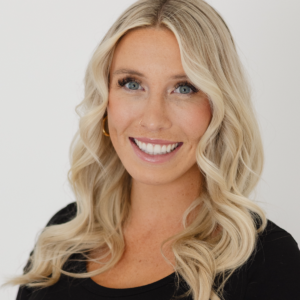 A woman with long, wavy blonde hair and blue eyes smiles at the camera. She is wearing a black top and a small nose ring, with a plain white background behind her.