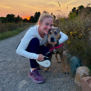 A smiling woman crouches beside a brown dog wearing a pink harness on a gravel path at sunset, with wildflowers and tall grass in the background.