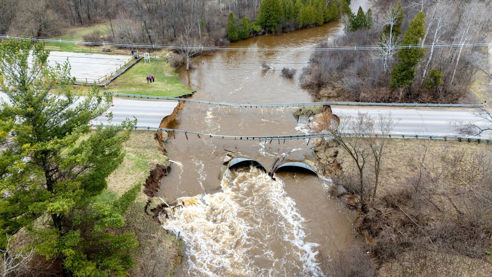 Aerial view of a road collapsed by flooding, with muddy water rushing through two culverts beneath the broken roadway, surrounded by trees and muddy banks.