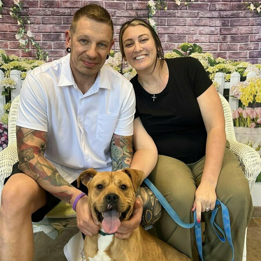 A smiling couple sits on a bench inside an adoption center, posing with a happy brown dog on a blue leash. The background features a floral wall and signs about pet adoption.