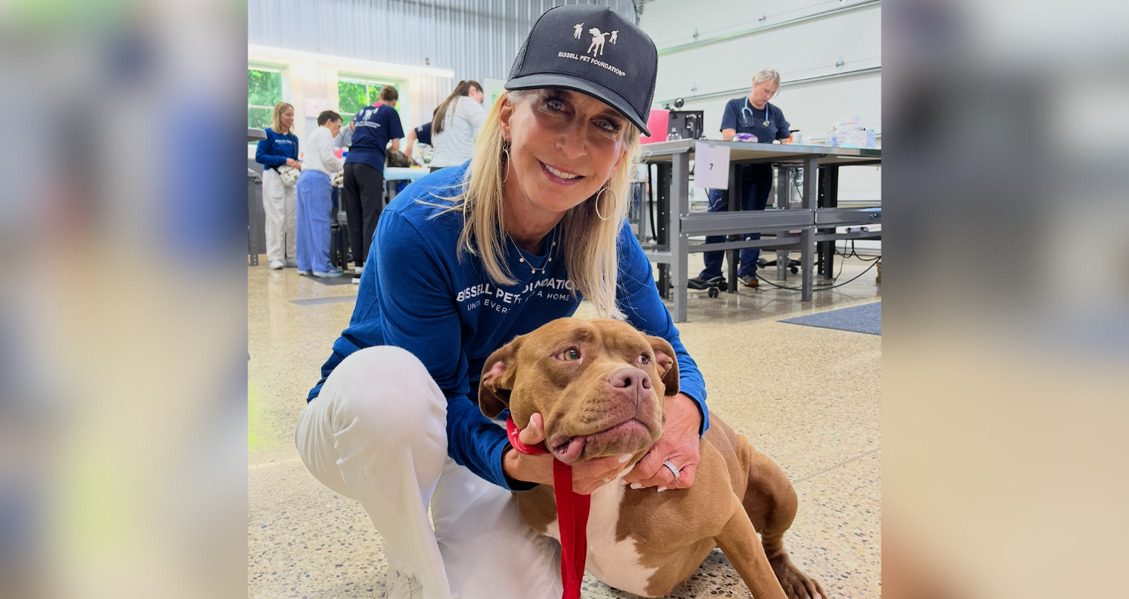 A woman in a black hat and blue shirt kneels indoors, smiling and hugging a brown dog with a red leash. Several people work at tables in the background of the bright, spacious room.
