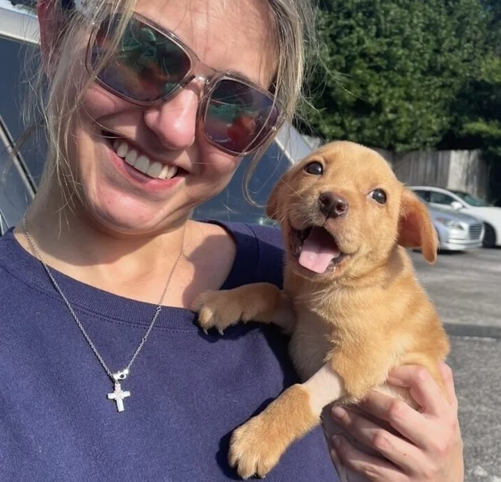 A smiling woman wearing sunglasses and a blue shirt holds a happy brown puppy outside in a parking lot on a sunny day.