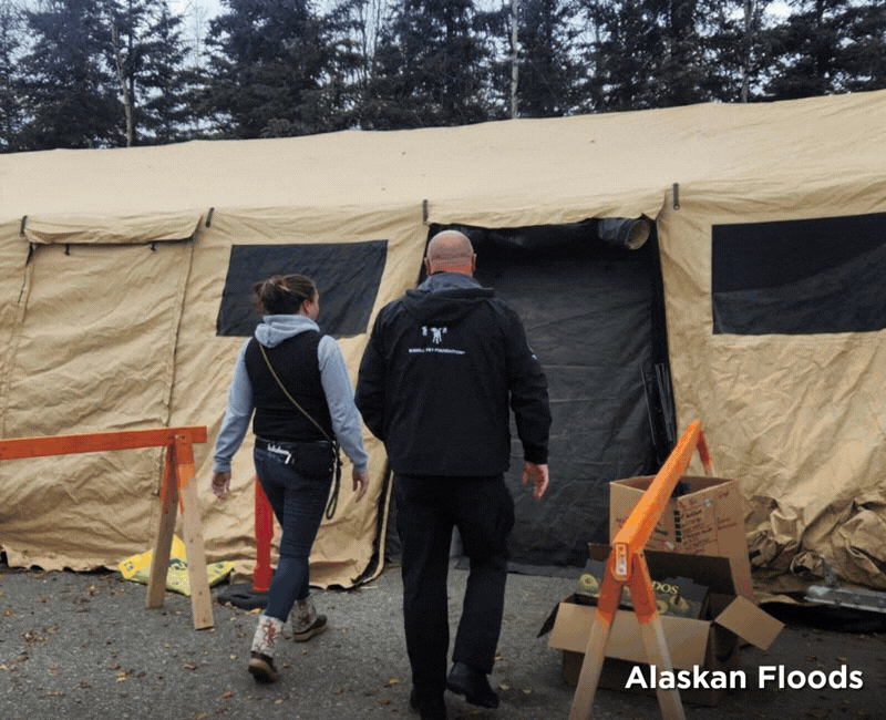 A woman and a man walk toward the entrance of a large tan emergency relief tent, with cardboard boxes and orange safety barriers nearby.