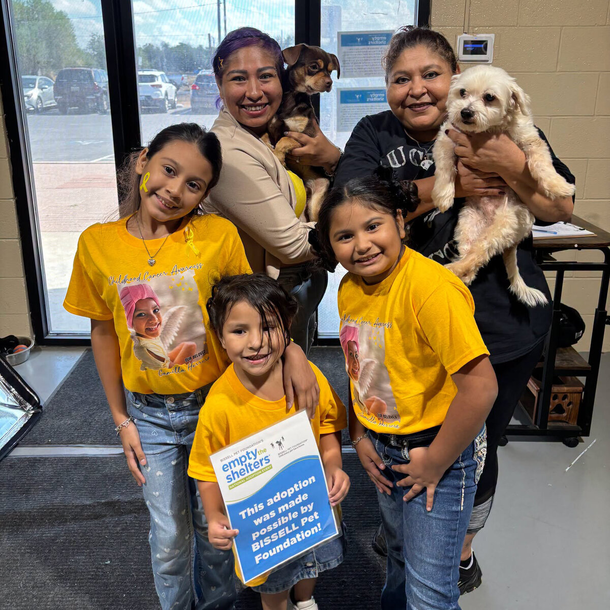 Two women and three young girls in matching yellow shirts pose indoors while holding two small dogs; the child in the front holds a sign about a pet adoption supported by the Bissell Pet Foundation.