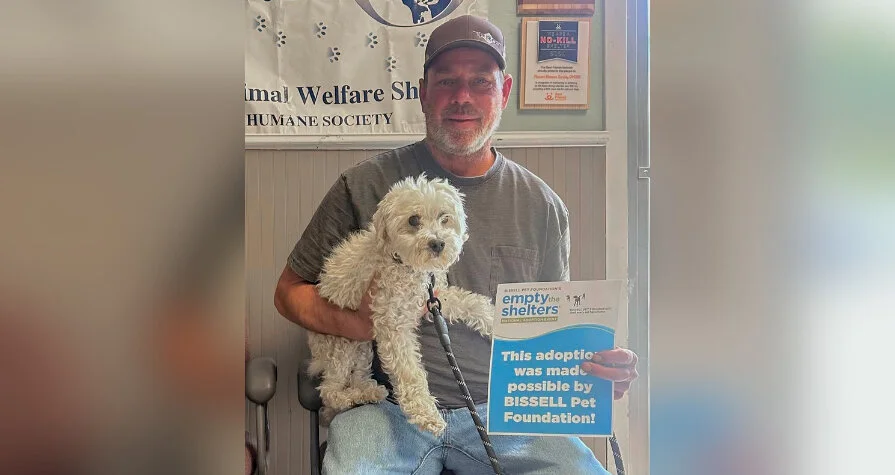 A man wearing a baseball cap holds a small white dog and a sign that reads, This adoption was made possible by BISSELL Pet Foundation!.